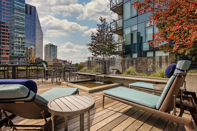 Rooftop lounge area with chaise lounges, tables, and a water feature, overlooking a cityscape.