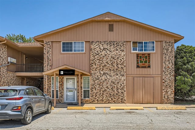 Exterior of a two-story apartment building with a front-office entrance, brick façade, and a parked car.