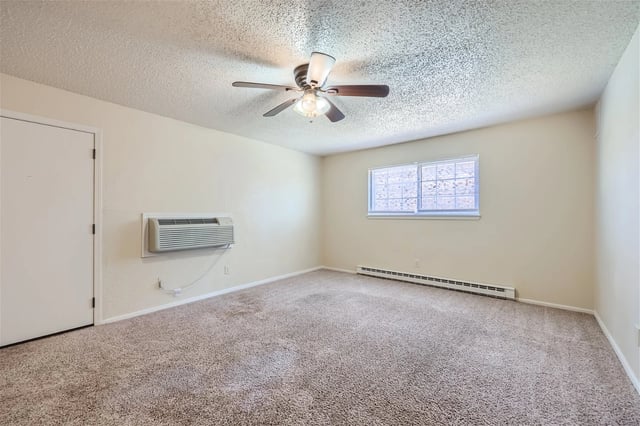 Vacant apartment room with beige walls, carpet, ceiling fan, window, wall AC unit, and baseboard heater.
