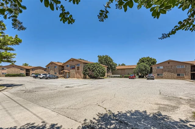 Exterior view of a brick apartment community with a parking lot and trees under a clear blue sky.