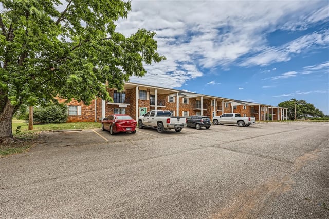 Apartment building exterior with a parking lot in front and a large tree on the left.