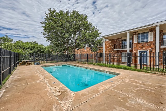 Swimming pool with blue water and concrete deck, next to a brick building and surrounded by a black fence.