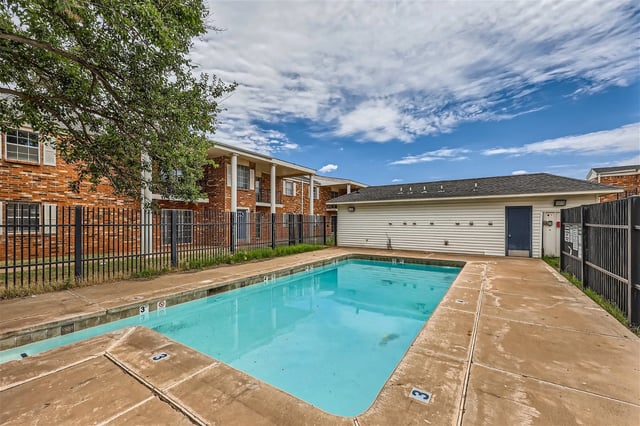 An inviting outdoor swimming pool with clear blue water, surrounded by a concrete deck and a black metal fence. Apartment buildings are visible in the background under a cloudy sky.