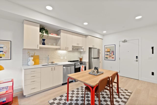 Open-concept kitchen in a modern apartment with light wood cabinets, stainless steel fridge, and wooden dining table.