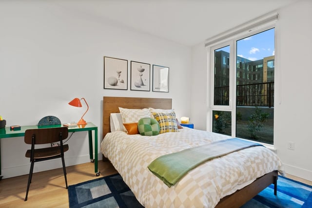 Bedroom in a modern apartment with a wooden bed, green desk, and a large window.