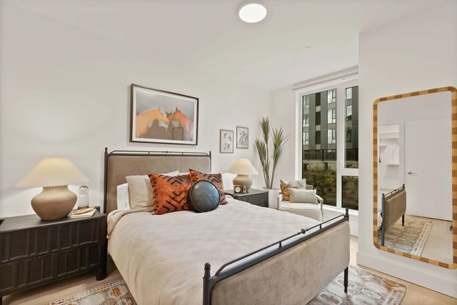 Bedroom in a modern apartment with a bed, nightstands, a large mirror, and a window view.