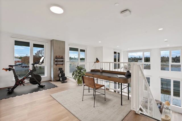 Loft-style community fitness area with a stationary bike, weights, and a desk near large windows.