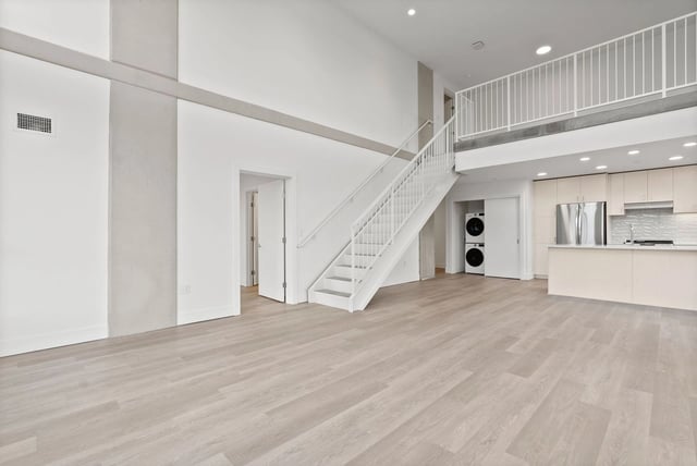 Open living area with light wood floors, white walls, a staircase to a loft, and a modern kitchen.