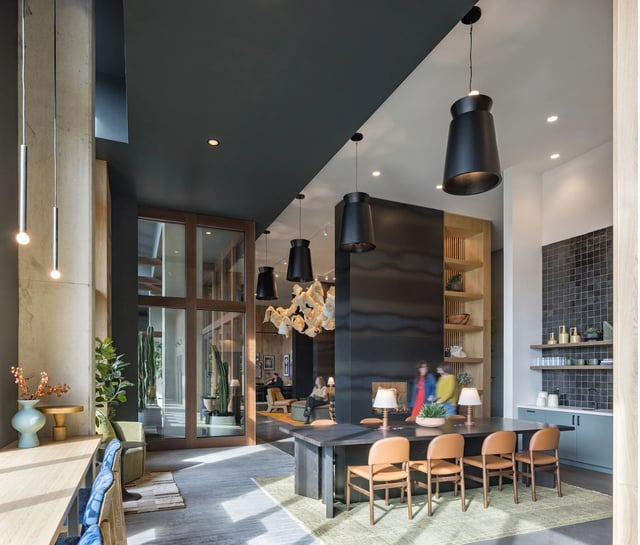 Modern community lounge with a long table, black pendant lights, and seating; people visible in the background.