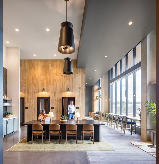 Modern apartment community lounge featuring a long table, seating, pendant lights, wood accent wall, and large windows.