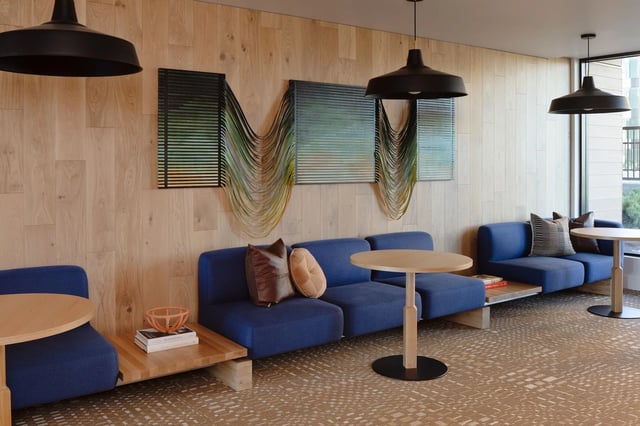Modern apartment community lounge with blue couches, wooden tables, wall art, and large windows.