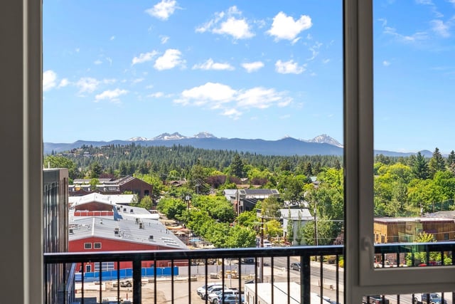 Balcony view of a town with trees and distant mountains under a blue sky.