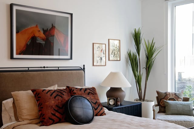 Bedroom with beige bedding, a metal bed frame, pillows, lamp, and tall plants by a window.
