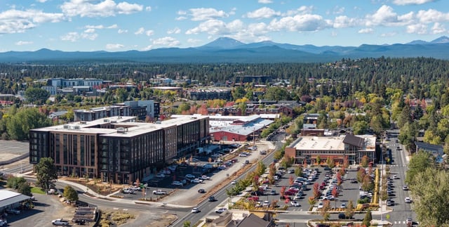 Aerial view of a multi-building apartment community with parking lots and surrounding trees, mountains in the distance.