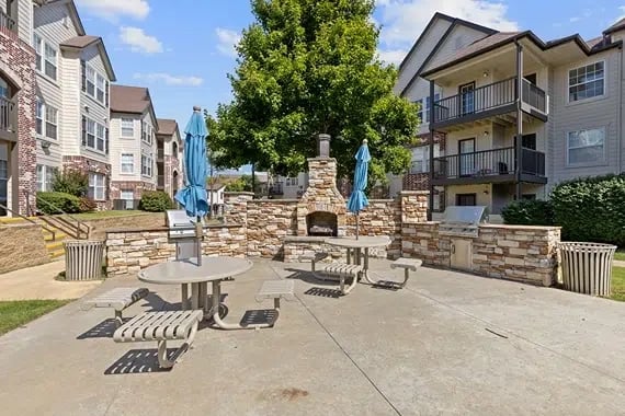 Outdoor communal courtyard with a stone fireplace, concrete dining tables, and blue umbrellas among apartment buildings.