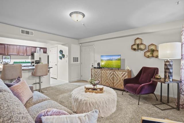 Living room in an apartment with a grey sofa, burgundy chair, TV on a wooden console, and an open kitchen.