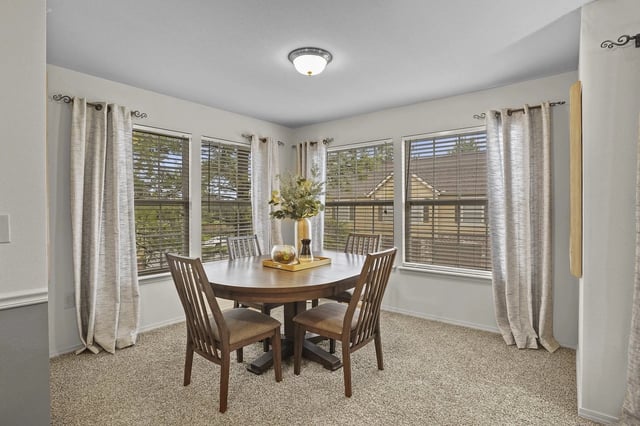 Dining room with a round table and four chairs, large windows with blinds, and decorative curtains.