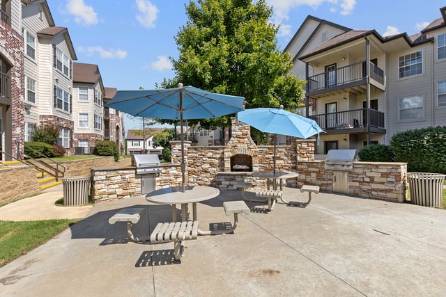 Outdoor seating area with picnic tables, umbrellas, grills, and a stone fireplace.