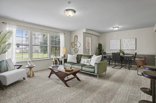 Bright apartment living room with a green sofa, white chairs, and large front windows.