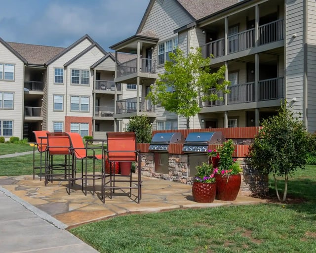Outdoor communal grilling area with orange chairs and built-in grills beside a multi-story apartment building.