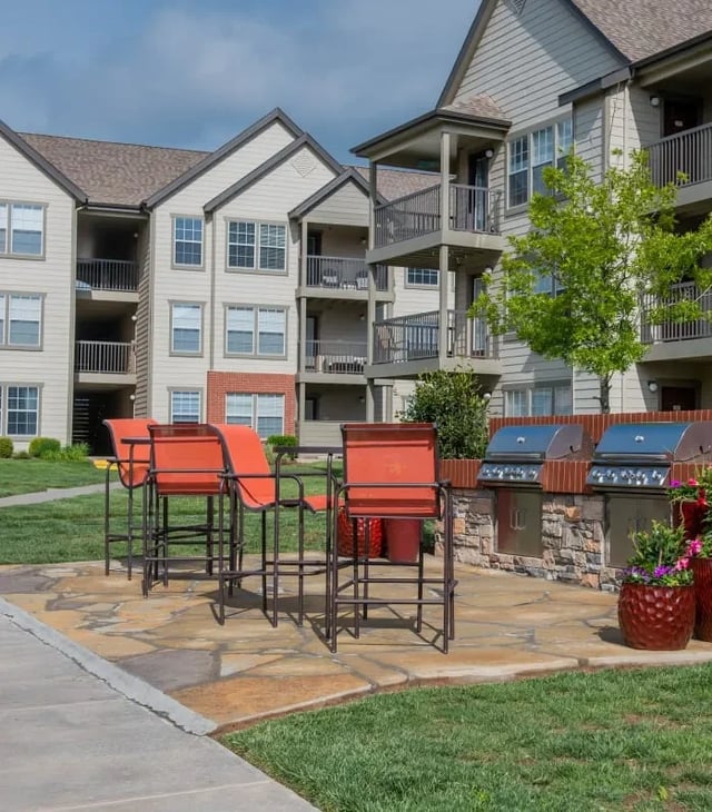 Outdoor community grilling area with red bar stools and built-in grills in front of a multi-story apartment.