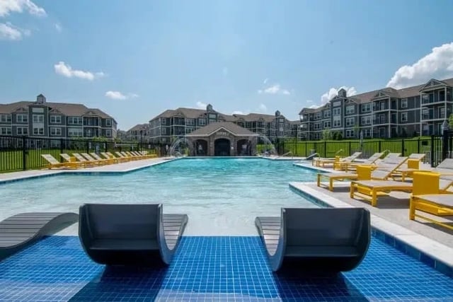 Outdoor community pool with blue-tiled deck, yellow lounge chairs, and apartment buildings in the background.
