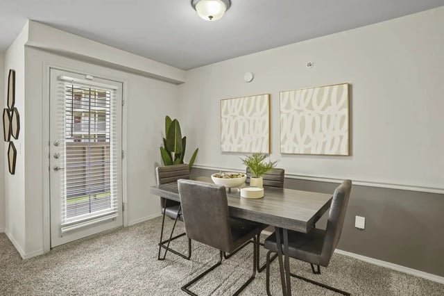 Dining area in a modern apartment with a gray table, chairs, plant, and wall art near a door with blinds.