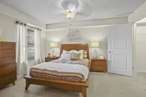 Furnished bedroom in an apartment featuring a wooden bed, two nightstands, lamps, a dresser, and a window with curtains.