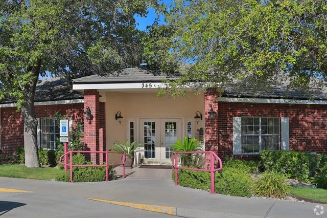 Building entrance with red brick facade and double glass doors, flanked by windows and lush greenery.