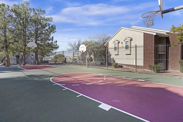 Outdoor basketball court with purple and green surface, fenced beside a brick building.