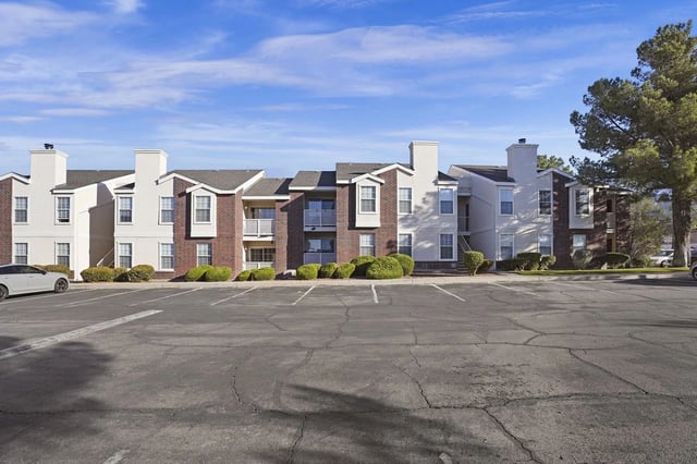 Exterior view of a brick-and-white apartment complex with a parking lot and manicured shrubs.