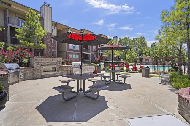 Outdoor gathering area with grills, picnic tables, umbrellas, and a pool in the background.