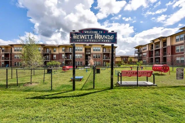 Exterior view of a pet-friendly community park with a fenced playground and apartment buildings in the background.