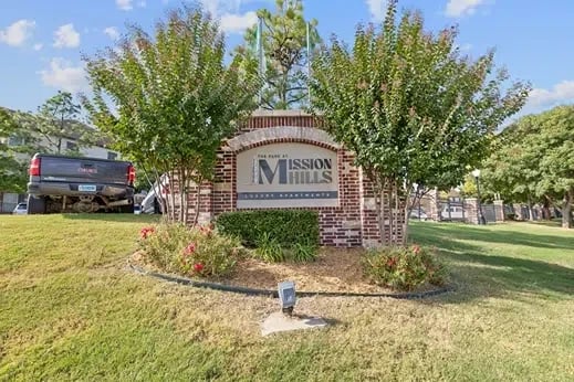Brick entrance sign for Mission Mills apartment community with trees and landscaped flower bed.