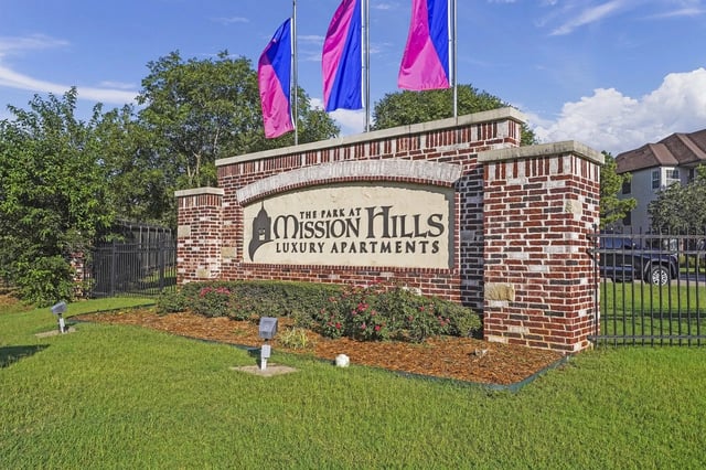 Brick entrance sign for The Park at Mission Hills Luxury Apartments with flagpoles and landscaping.