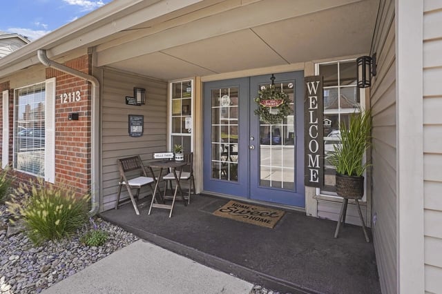 Front entrance of an apartment building with blue double doors, a welcome sign, and a small seating area.