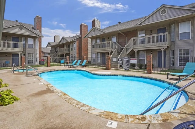 Outdoor community pool at an apartment complex with lounge chairs and surrounding buildings.