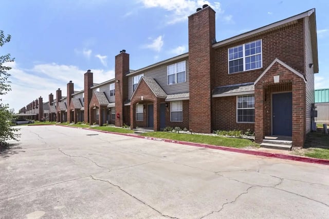 Exterior view of brick townhouse-style apartment buildings arranged in a row along a paved parking area.
