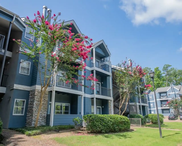 Exterior view of a blue multi-story apartment building with balconies and pink flowering trees.