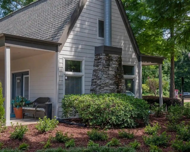 Exterior of a small apartment building with gray siding, stone chimney, and landscaped front yard.