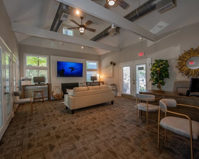 Interior communal lounge with beige sofa, desks, a wall-mounted TV, ceiling fans, and exposed ducts.