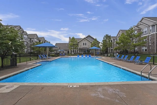 Outdoor apartment community pool with blue lounge chairs and umbrellas, surrounded by buildings.