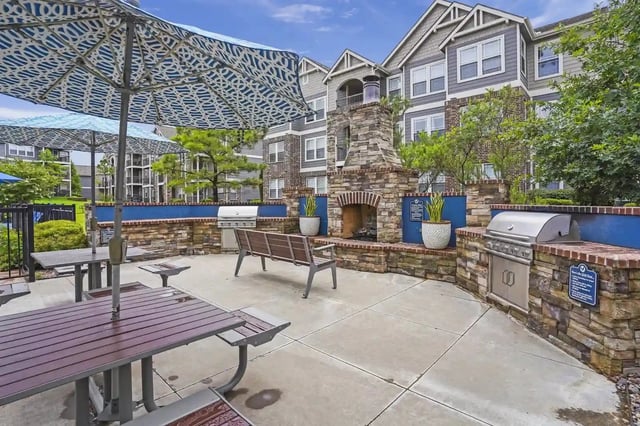 Outdoor community amenity area with a stone fireplace, grills, benches, and umbrellas.