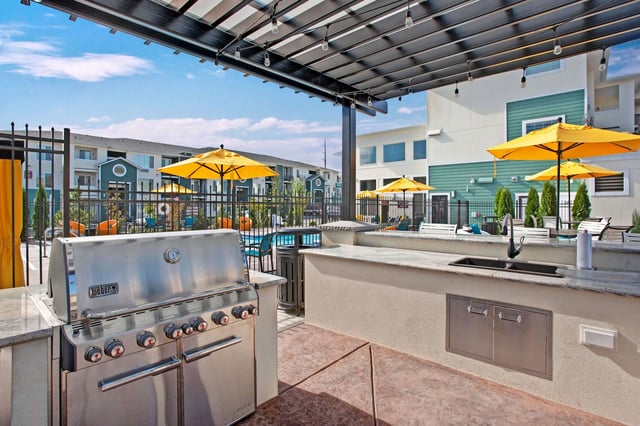 Outdoor grilling station with a stainless steel grill, sink, and granite countertop, next to a swimming pool area with yellow umbrellas.