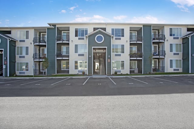 Exterior view of a modern apartment building with balconies and a parking lot in the foreground.