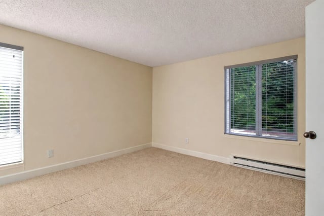 Empty apartment bedroom with beige walls and carpet, featuring two windows with blinds.