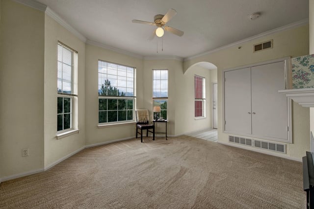 Spacious living room with large windows overlooking trees and mountains, featuring carpet flooring and a ceiling fan.