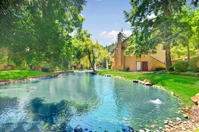 Apartment buildings bordering a serene pond with fountains and lush greenery.