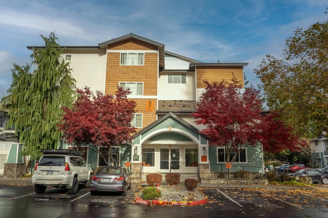 Exterior of an apartment building with red and orange trees and a parking lot in front.