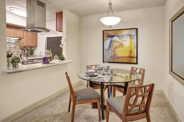 Dining area with a glass-top table and chairs, adjacent to a kitchen pass-through.